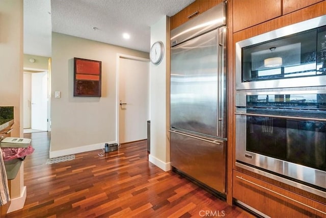 kitchen with dark wood-type flooring, baseboards, built in appliances, brown cabinets, and a textured ceiling