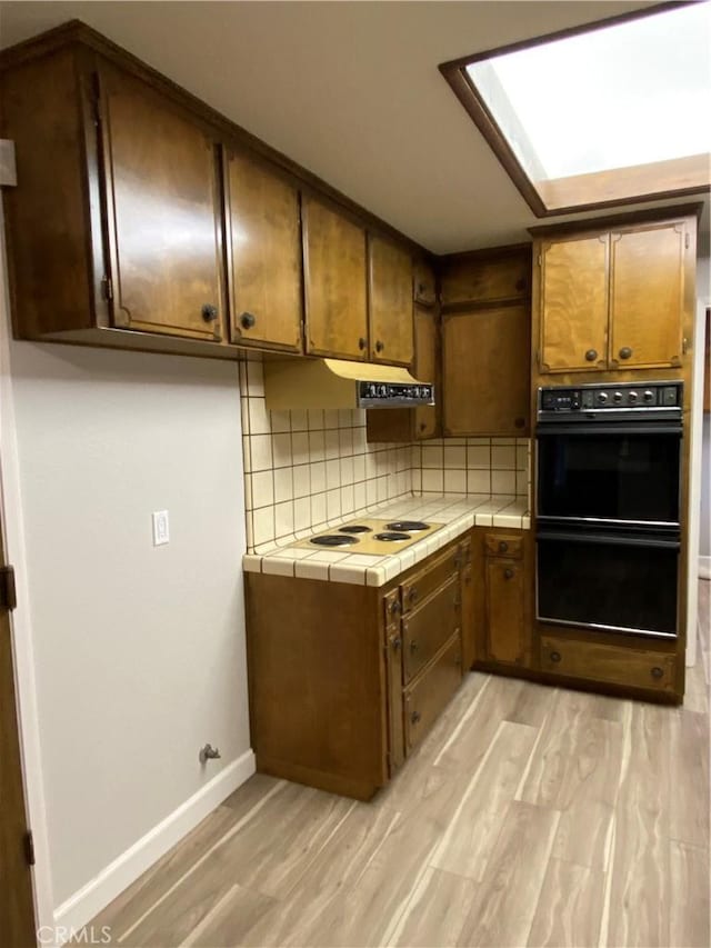 kitchen with light wood-style flooring, dobule oven black, tasteful backsplash, white electric cooktop, and tile counters