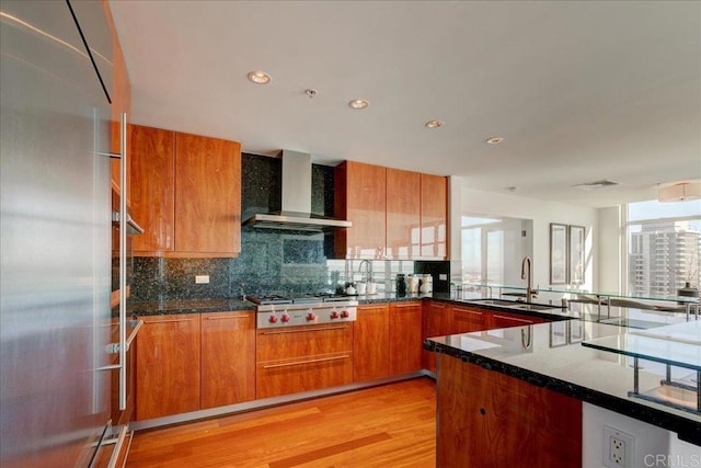 kitchen featuring a healthy amount of sunlight, appliances with stainless steel finishes, brown cabinetry, wall chimney exhaust hood, and a sink