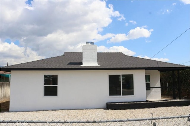 back of house with stucco siding, fence, a chimney, and a shingled roof