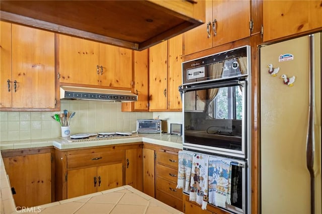 kitchen featuring under cabinet range hood, decorative backsplash, tile countertops, and freestanding refrigerator