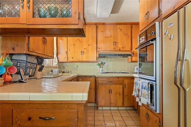 kitchen with tasteful backsplash, tile counters, under cabinet range hood, light tile patterned floors, and freestanding refrigerator