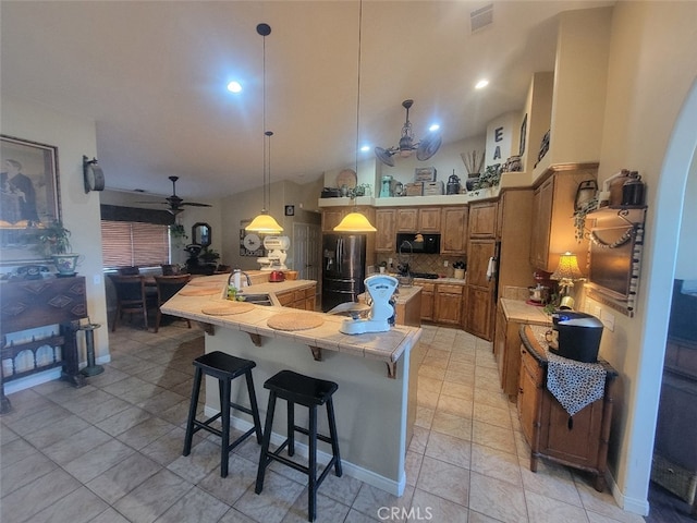 kitchen with black appliances, tile countertops, brown cabinetry, ceiling fan, and decorative backsplash