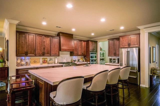 kitchen featuring dark wood-style flooring, appliances with stainless steel finishes, an island with sink, ornamental molding, and custom exhaust hood