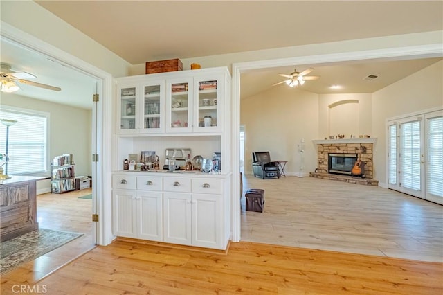 interior space with light wood-type flooring, a stone fireplace, a ceiling fan, and white cabinetry