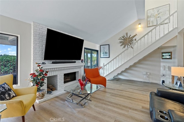 living area with stairs, high vaulted ceiling, a brick fireplace, and wood finished floors