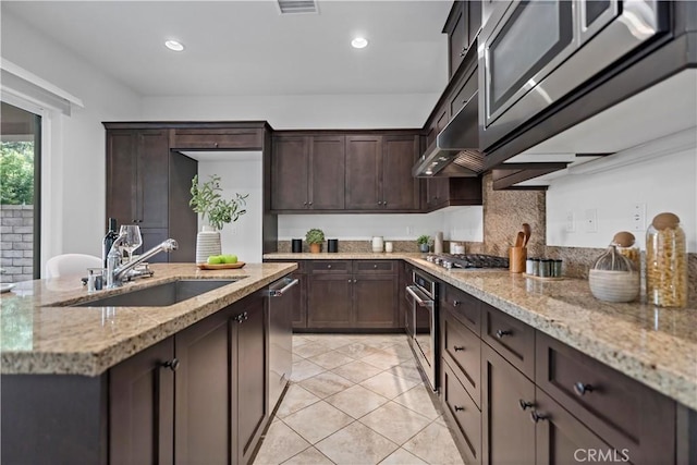 kitchen with dark brown cabinets, appliances with stainless steel finishes, under cabinet range hood, a sink, and light stone counters