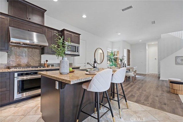 kitchen with visible vents, a kitchen island with sink, appliances with stainless steel finishes, ventilation hood, and light tile patterned floors