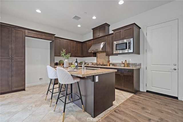 kitchen featuring a sink, range hood, stainless steel microwave, a kitchen island with sink, and visible vents