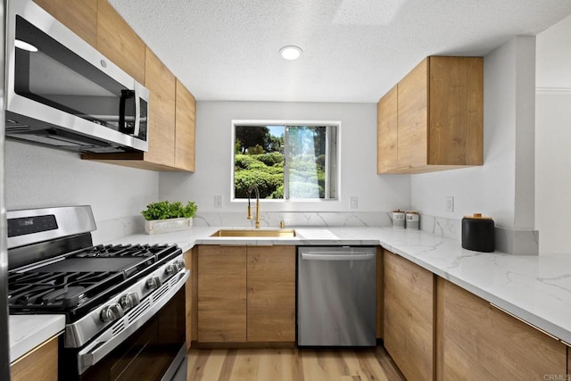 kitchen featuring a sink, stainless steel appliances, light wood-style floors, brown cabinetry, and light stone countertops