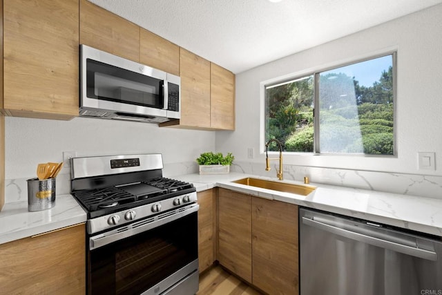 kitchen with a sink, a textured ceiling, stainless steel appliances, brown cabinetry, and light stone countertops