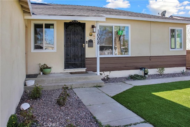 entrance to property with stucco siding, roof with shingles, covered porch, and a yard