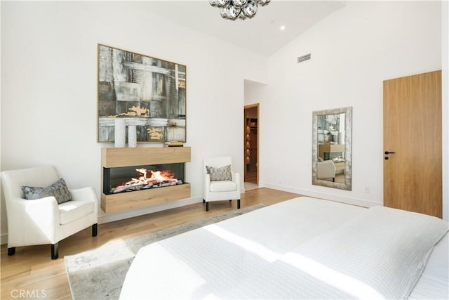 bedroom featuring wood finished floors, visible vents, high vaulted ceiling, a warm lit fireplace, and a notable chandelier