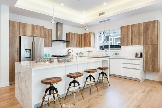 kitchen with a tray ceiling, modern cabinets, wall chimney exhaust hood, and stainless steel refrigerator with ice dispenser