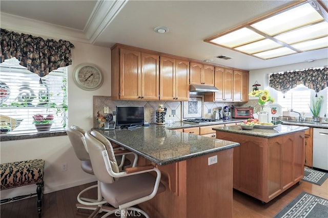 kitchen with ornamental molding, under cabinet range hood, dark stone countertops, tasteful backsplash, and gas stovetop