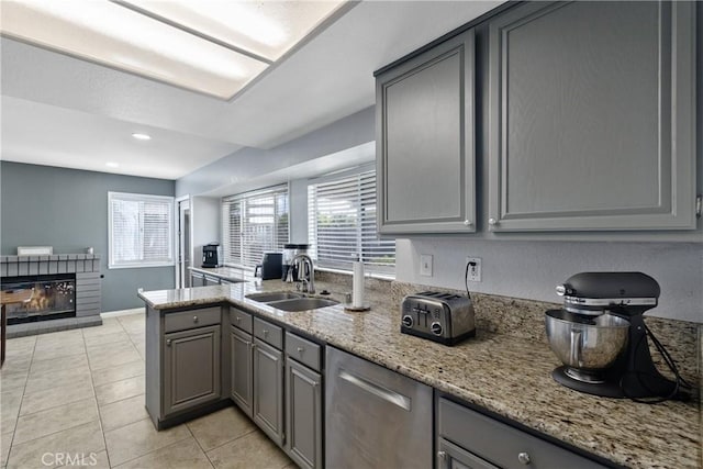 kitchen featuring gray cabinets, stainless steel dishwasher, a brick fireplace, a sink, and light tile patterned flooring