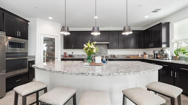 kitchen featuring appliances with stainless steel finishes, a breakfast bar area, an island with sink, light stone counters, and decorative light fixtures