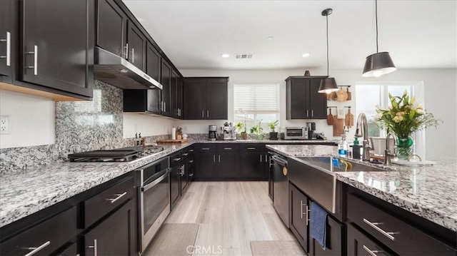 kitchen with under cabinet range hood, stainless steel appliances, a sink, light wood-style floors, and light stone countertops