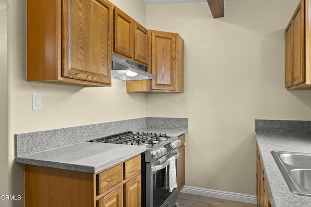 kitchen featuring baseboards, stainless steel range with gas stovetop, a sink, under cabinet range hood, and brown cabinets