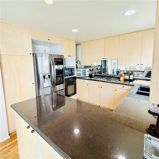 kitchen featuring light brown cabinetry, a sink, stainless steel appliances, dark stone counters, and a peninsula