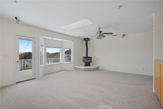 unfurnished living room with carpet, a skylight, baseboards, ceiling fan, and a wood stove