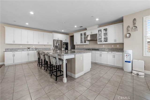 kitchen featuring under cabinet range hood, an island with sink, light tile patterned flooring, stainless steel appliances, and white cabinetry