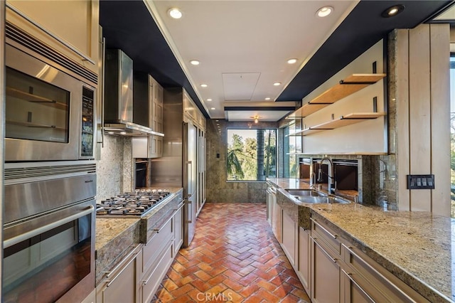 kitchen with open shelves, wall chimney range hood, light stone counters, and a sink