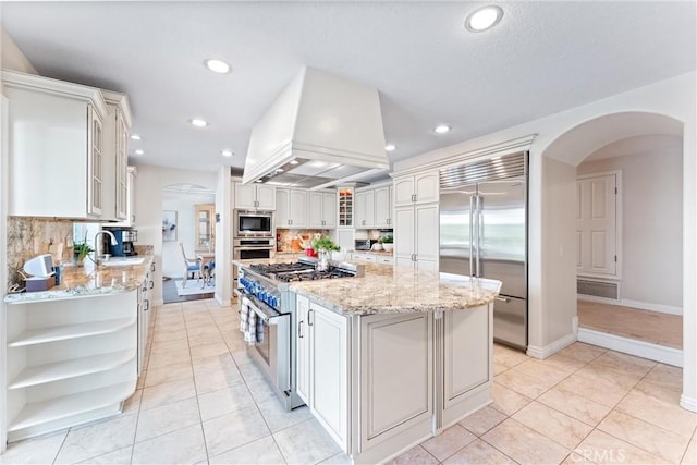 kitchen with built in appliances, custom exhaust hood, open shelves, arched walkways, and a sink