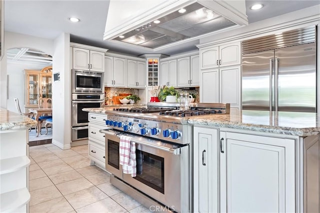 kitchen with built in appliances, premium range hood, arched walkways, tasteful backsplash, and light stone countertops
