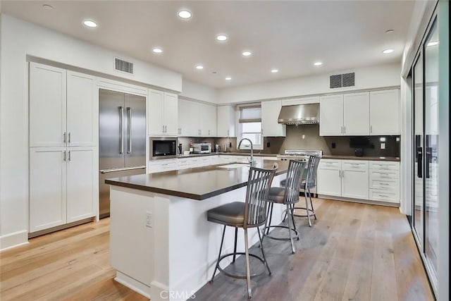 kitchen with wall chimney range hood, light wood-style floors, stainless steel appliances, white cabinetry, and visible vents