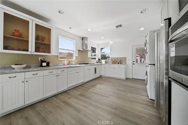 kitchen with white cabinetry, stacked washing maching and dryer, wall chimney exhaust hood, decorative backsplash, and light wood finished floors