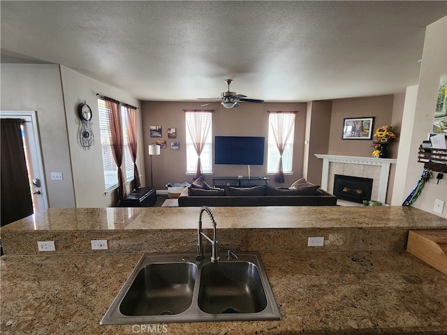 kitchen with a textured ceiling, tile counters, a tiled fireplace, a ceiling fan, and a sink