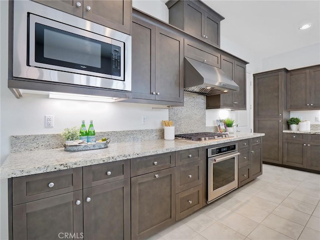 kitchen featuring light stone countertops, stainless steel appliances, dark brown cabinetry, under cabinet range hood, and backsplash
