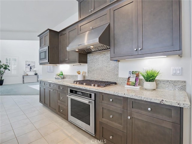 kitchen with tasteful backsplash, stainless steel appliances, range hood, light stone counters, and dark brown cabinetry