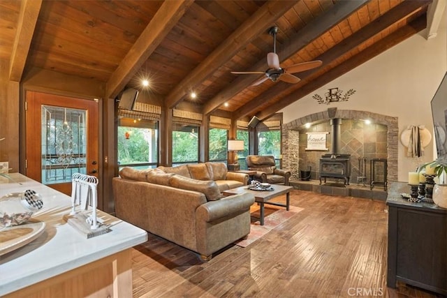 living area featuring vaulted ceiling with beams, ceiling fan, wood ceiling, a wood stove, and wood finished floors