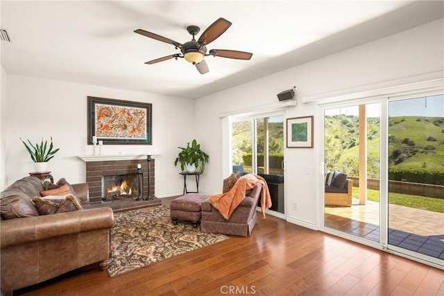 living area with a ceiling fan, hardwood / wood-style floors, baseboards, a brick fireplace, and visible vents