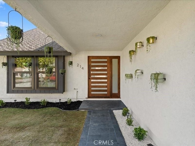 entrance to property with a shingled roof, a yard, and stucco siding