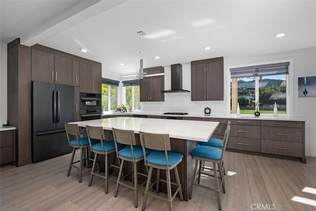 kitchen featuring wall chimney range hood, black appliances, a breakfast bar, a center island, and light wood finished floors