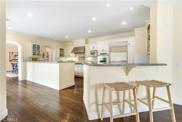 129 Via Waziers, Newport Beach, CA 92663 - kitchen featuring a peninsula, wall chimney range hood, appliances with stainless steel finishes, arched walkways, and glass insert cabinets