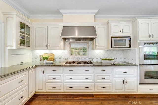 129 Via Waziers, Newport Beach, CA 92663 - kitchen featuring appliances with stainless steel finishes, wall chimney exhaust hood, dark stone counters, glass insert cabinets, and backsplash