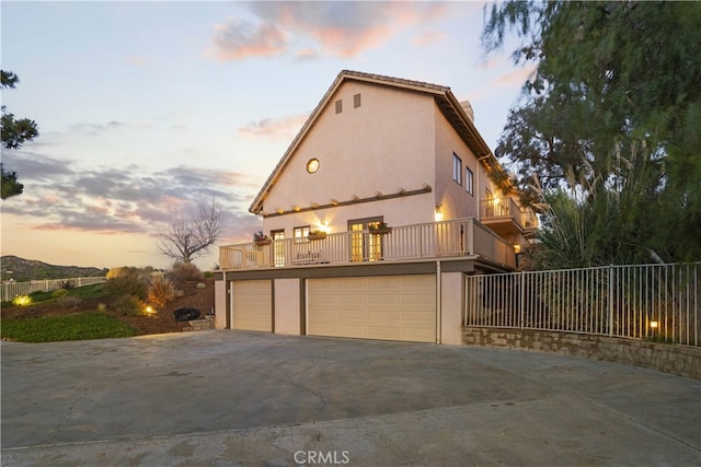 property exterior at dusk with an attached garage, a balcony, driveway, and stucco siding