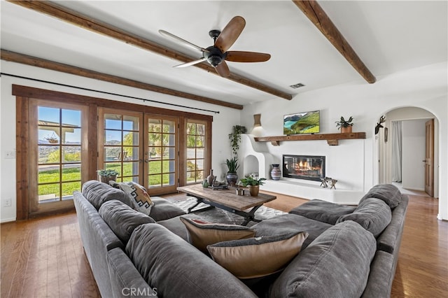living area featuring beamed ceiling, arched walkways, a ceiling fan, visible vents, and wood finished floors