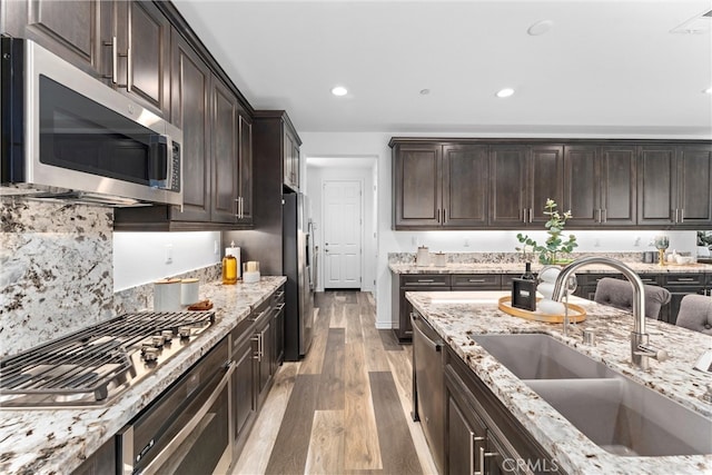 kitchen with light stone counters, dark brown cabinets, a sink, stainless steel appliances, and light wood finished floors