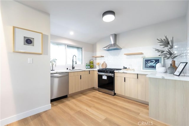 kitchen featuring gas range oven, light brown cabinetry, stainless steel dishwasher, a sink, and wall chimney range hood