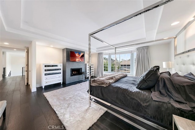 bedroom with a warm lit fireplace, a tray ceiling, recessed lighting, and dark wood-style floors