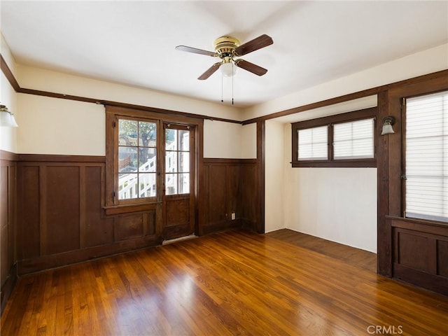 unfurnished room with wood finished floors, plenty of natural light, a wainscoted wall, and a ceiling fan