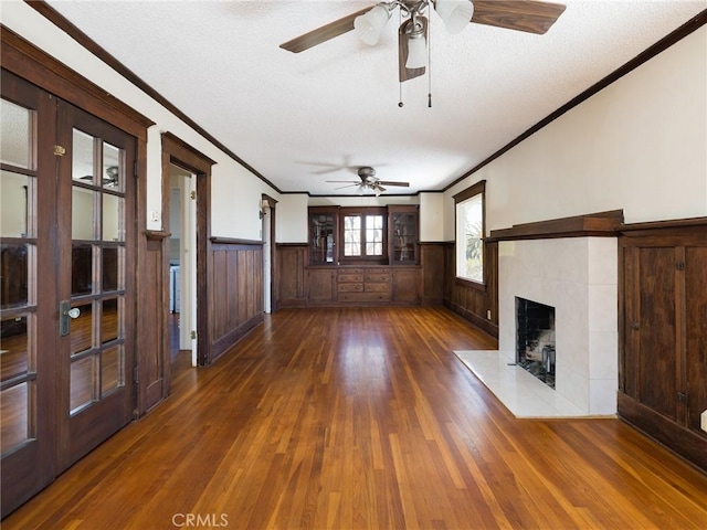 unfurnished living room with ceiling fan, a wainscoted wall, ornamental molding, a fireplace, and wood finished floors