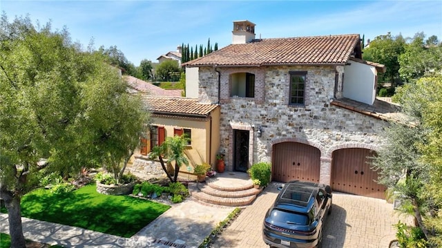 mediterranean / spanish-style house with a tiled roof, stone siding, a garage, a chimney, and decorative driveway