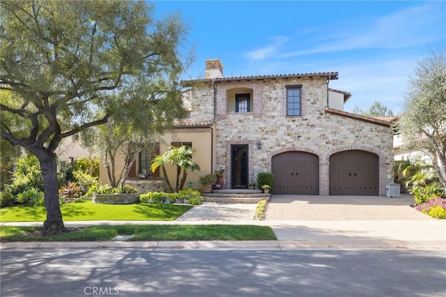 mediterranean / spanish-style house featuring decorative driveway, stone siding, a front lawn, a chimney, and a garage