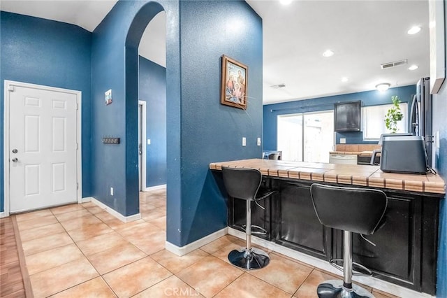 kitchen featuring visible vents, light tile patterned flooring, arched walkways, tile counters, and a kitchen breakfast bar